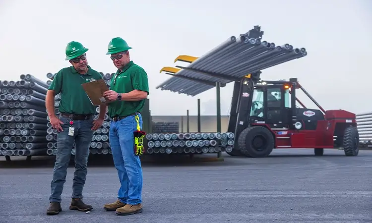 Two Workstrings workers looking over a clipboard while a forklift moves drill pipes in the background