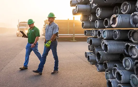 Two Workstrings workers walking by a rack of heavy drill strings at sunrise 