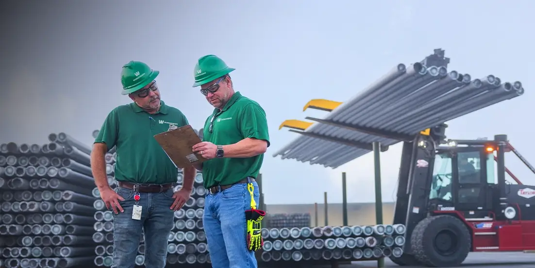 Two Workstring workers looking over inventory details on a clipboard while a forklift moves drill pipes behind them