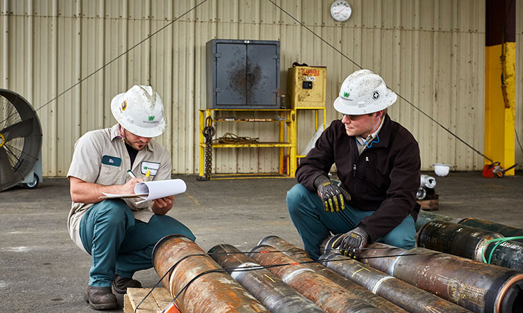 Two Workstrings workers kneeling down looking over pipes and writing down notes on a clipboard